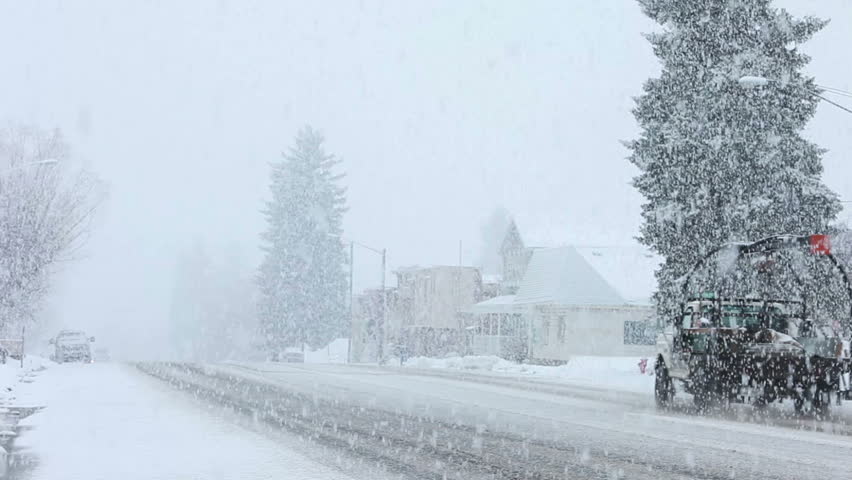 Truck and car drives through heavy snow blizzard in late winter on main street of a small country town. City center with old pioneer businesses.