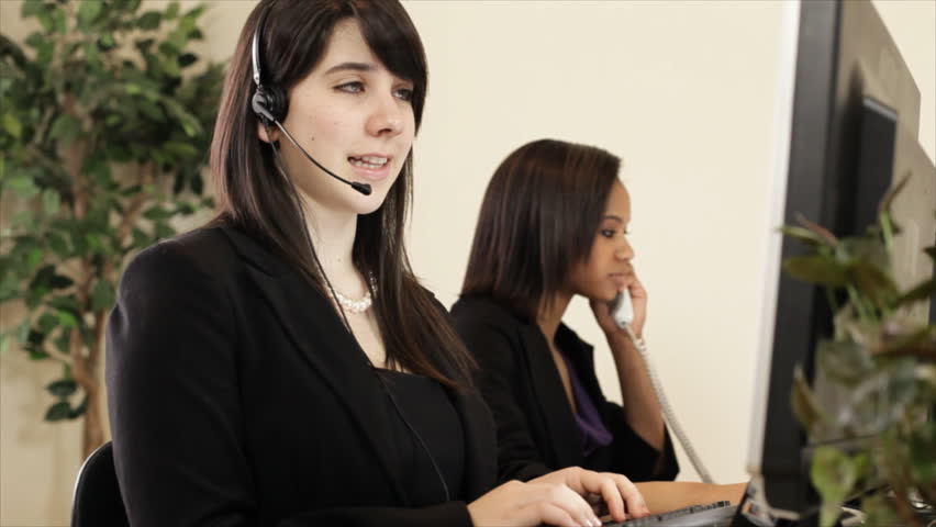 A lovely girl and her pretty African American co-worker working the helpdesk using a headset and a regular desk phone. Shallow depth of field.
