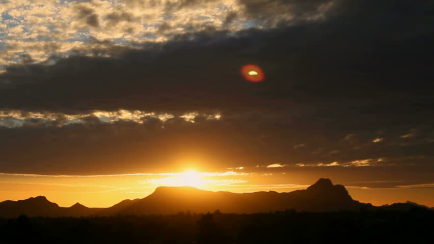 Time Lapse, Sunlight seeps through clouds, projecting what looks like second fiery yellow-orange sun into sky as sun sets over mountains, slowed. 1080p