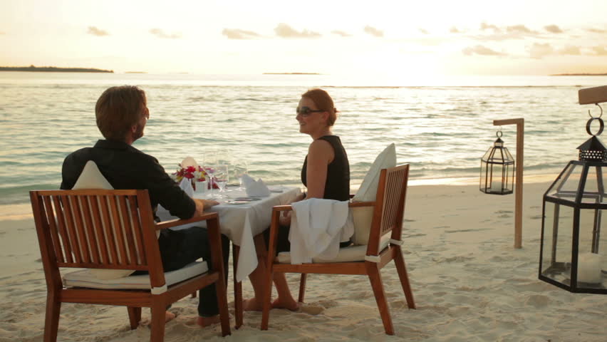 young couple sitting and dining at a tropical beach in front of the ocean at sunset with audio 