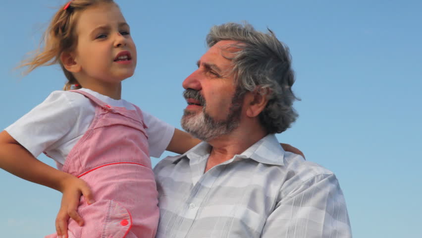 elderly man with his granddaughter on hands,he something shows and tells her against sky in summer 