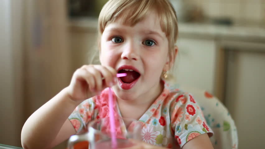 Little girl drinking from a glass of juice through a straw. Laughs. Looks at camera