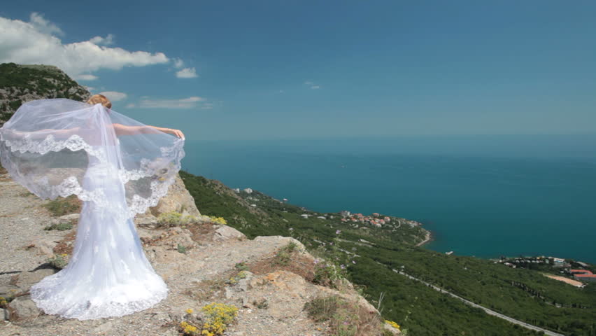 bride stands on cliff with a flowing veil on the wind against the blue sea and sky
