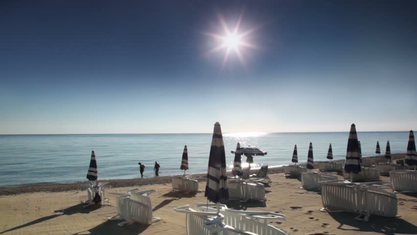 Many parasols and deckchairs standing on beach at sunny day, two tourists stand in water and woman looking something in water