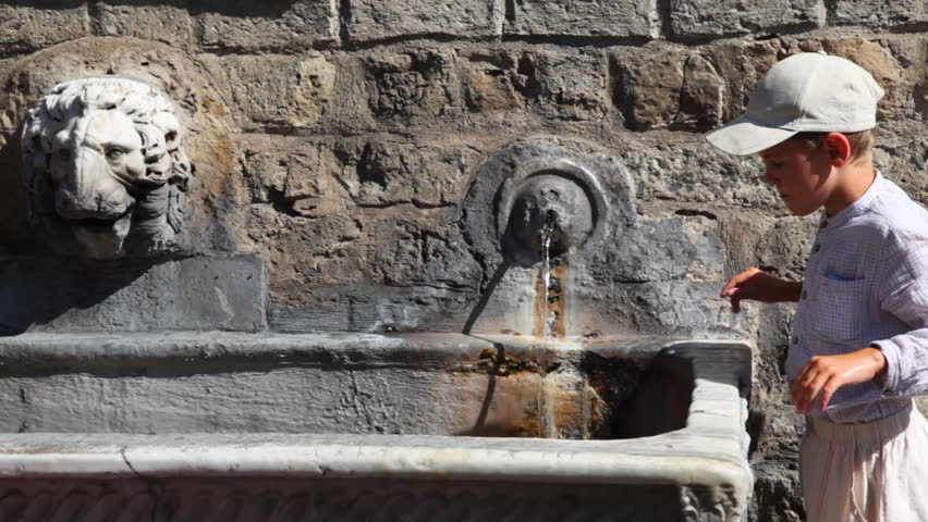little boy stands and drinks water from old source on stone wall in sunny day