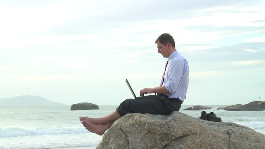 Businessman working with laptop while sitting on a rock on the beach.