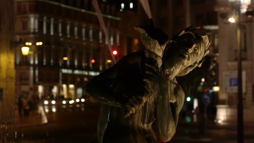 Night time video of a fountain statue with cars and people passing in the background.