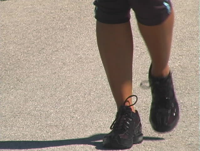 A beautiful woman walking along a jogging track outdoors.  Shot begins with a close-up of her feet and tilts up.