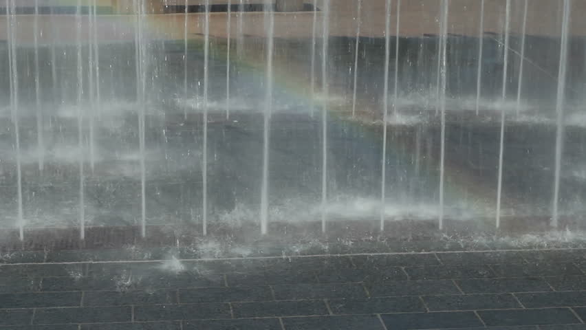 Water jets spraying H20 up out of paver bricks on a bright sunny day causing a rainbow to appear in the water mist in a public park.