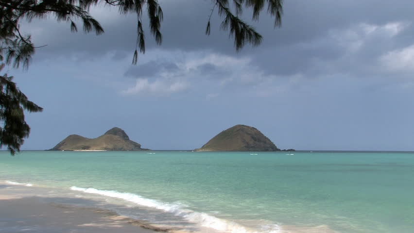 Tropical beach on Oahu, Hawaii. Islands off shore in Pacific ocean. Two outriggers paddling by distance islands. Waves on white sand with tree limbs overhead. Scenic, relaxing, calm seascape.