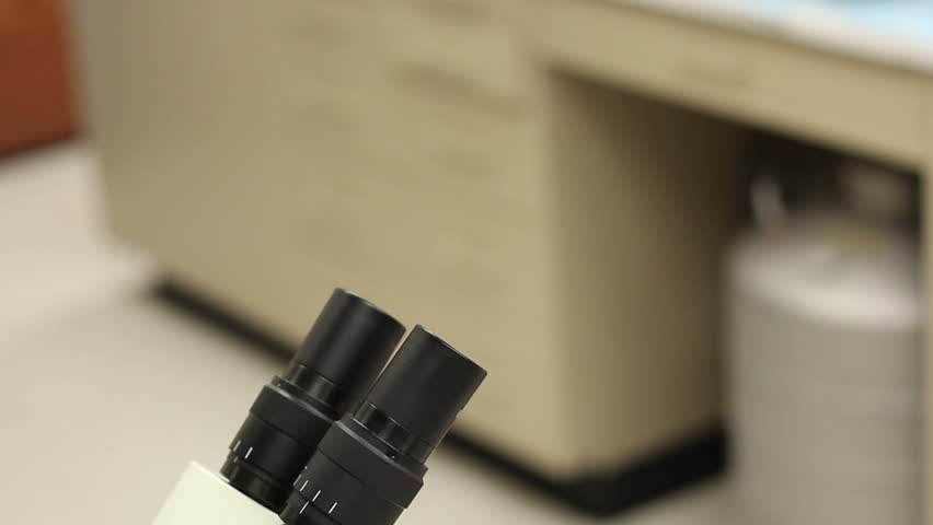 Asian researcher in a labcoat and goggles looks into a microscope.