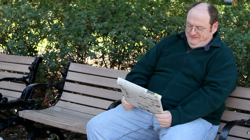 Overweight man sits on a park bench reading the newspaper.