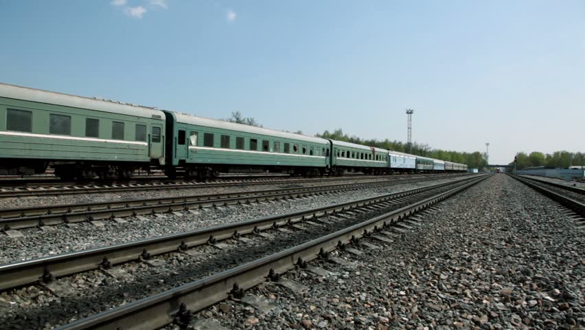 Short train passes by on railway, other stands on parallel rails, traffic on bridge far