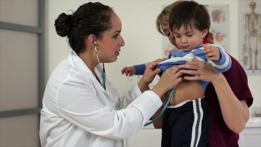 A nurse holds a cute little playful toddler while a pretty Hispanic pediatrician tries to listen to his breathing.