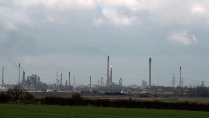 Industrial chimneys belching out smoke into the sky with traffic on the motorway in the foreground.