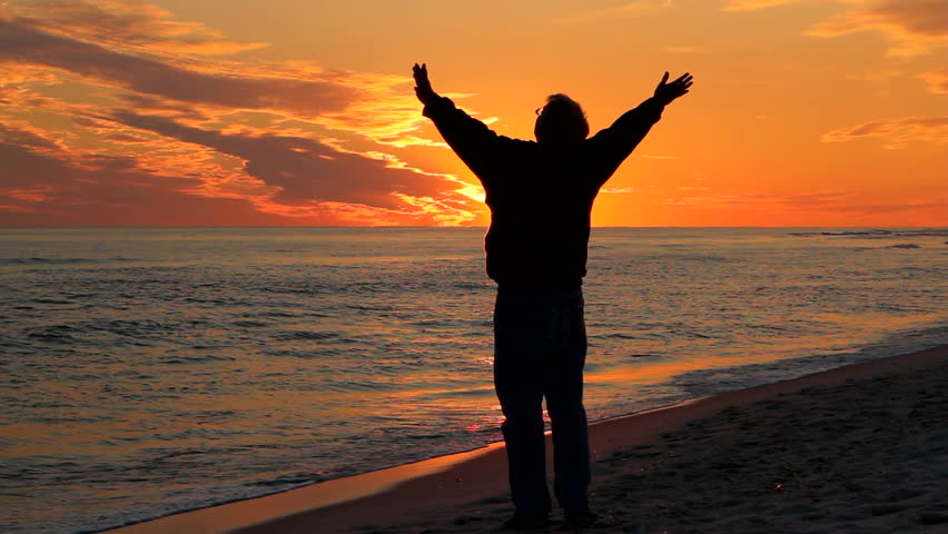 Man, with arms raised toward the heavens, prays at the beach at sunset.