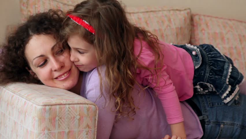Little girl lies on mother legs and kisses her cheek, closeup view at sofa