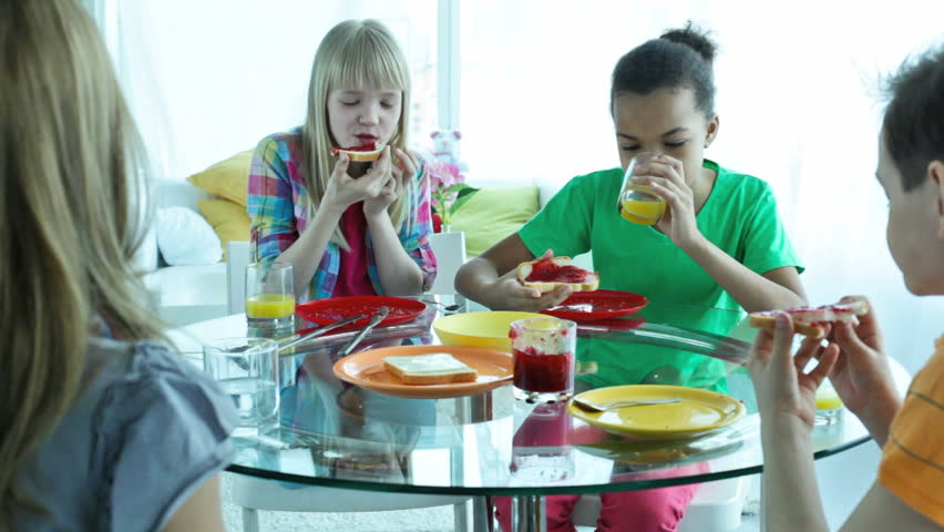 Cute kids sitting at table, having a snack and talking to a woman viewed from behind