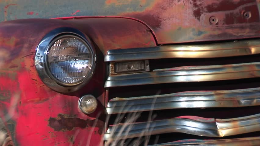 Close-up of dented and rusting abandoned pickup truck showing front passenger side headlight, grill, and hood.