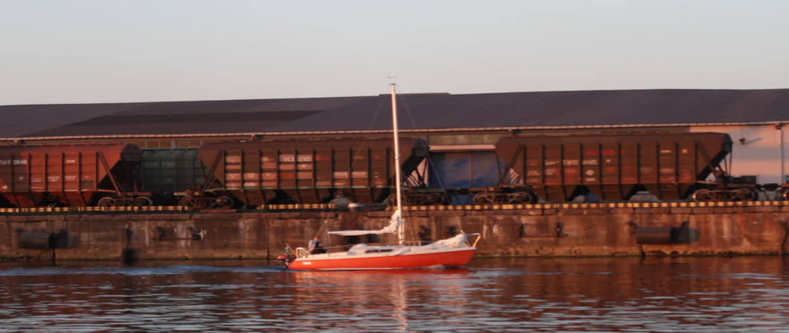 Sailboat moving at the Riga Freeport along the Daugava river, in Riga, Latvia. Port crane, container terminal and storage facility in the background.