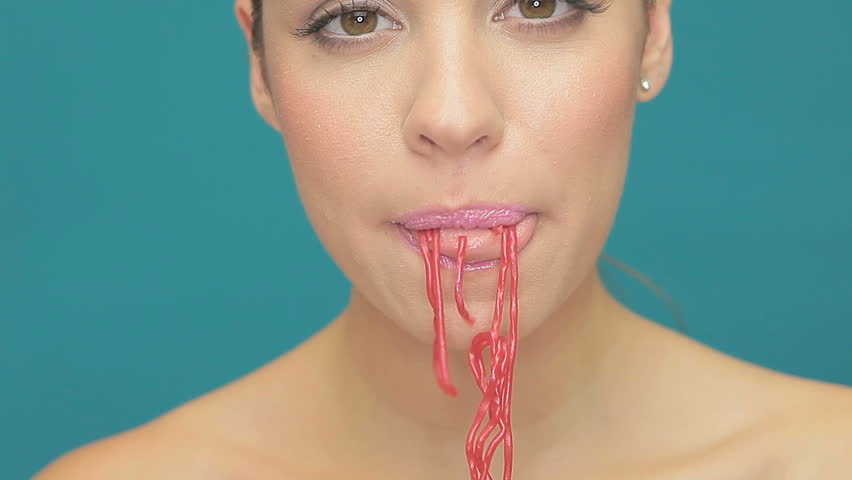 Closeup of the face of a sensual beautiful young woman eating long red strands of jelly sweets on a blue studio background