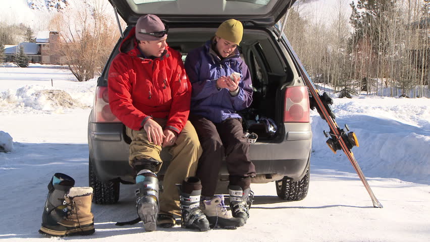 Couple sitting at the back of car talking on cell phone and putting on ski boots