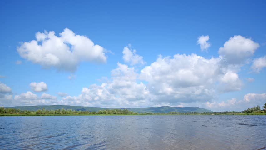 Clouds fly on blue sky over river in sunny weather, time lapse