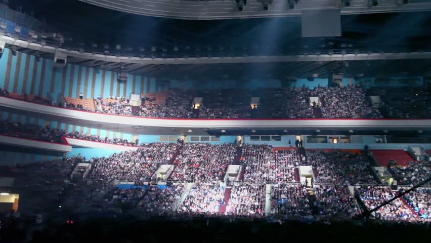 Searchlights in huge sports complex with lot of people on tribunes at concert, panoramic motion