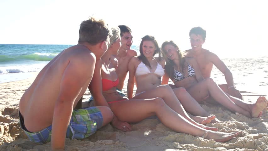 Group of teenage friends sitting on beach in swimsuits talking and laughing.