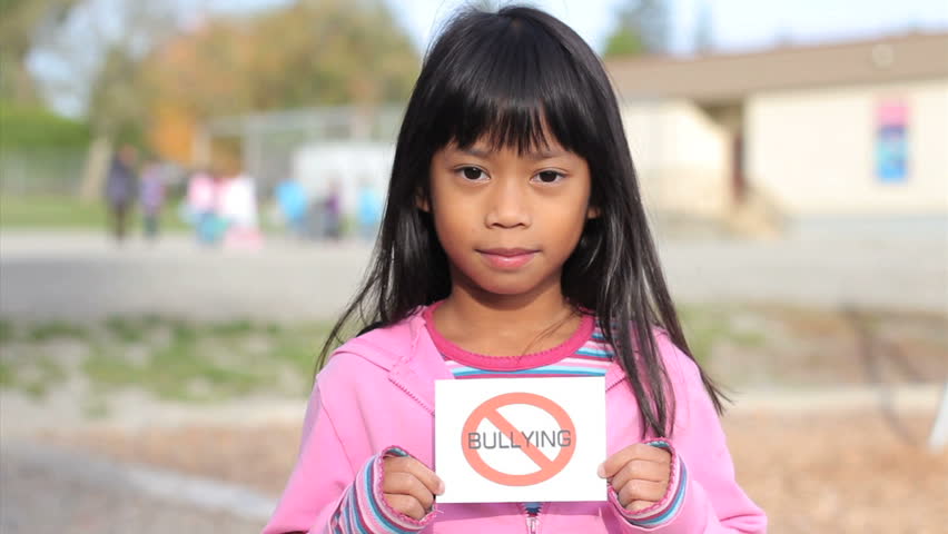 A cute Asian girl holds up a small NO BULLYING sign on the school playground.