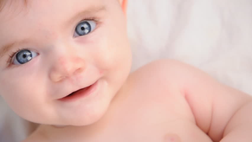 A cute little baby is looking into the camera and is happy on a white bed sheet. The baby looks around and then smiles and laughs.