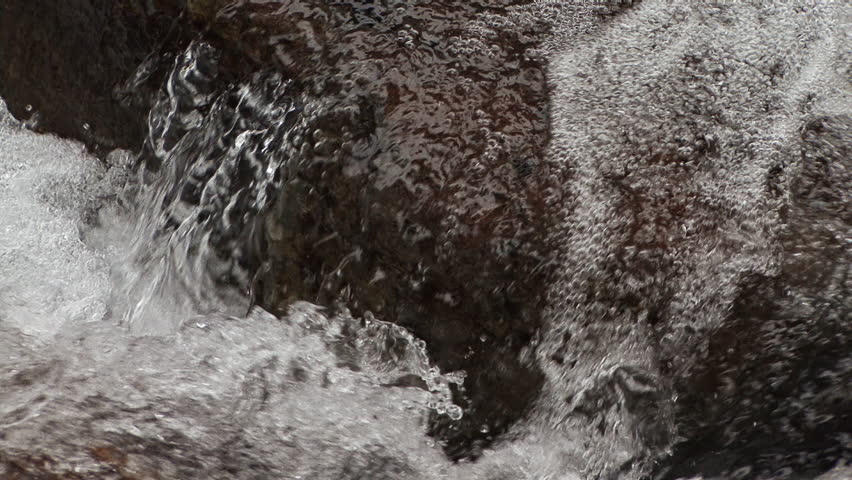 Incredibly clear water from high altitude snowmelt rushes down a small creek over reddish-hued rock, bubbling and splashing wildly in this extreme close-up shot.