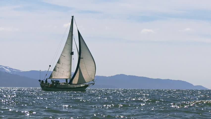A sailboat, either cutter or masthead sloop, under way across the glistening waves of Kachemak Bay in Homer, Alaska, as seen from the Homer Spit beach inches above sea level.