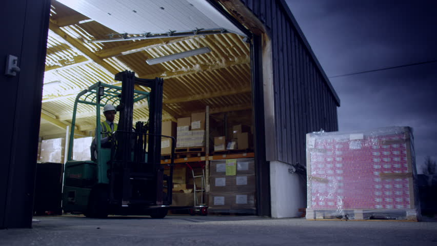 One warehouse worker drives a forklift truck out of a warehouse at night. His co-worker follows him out on foot.