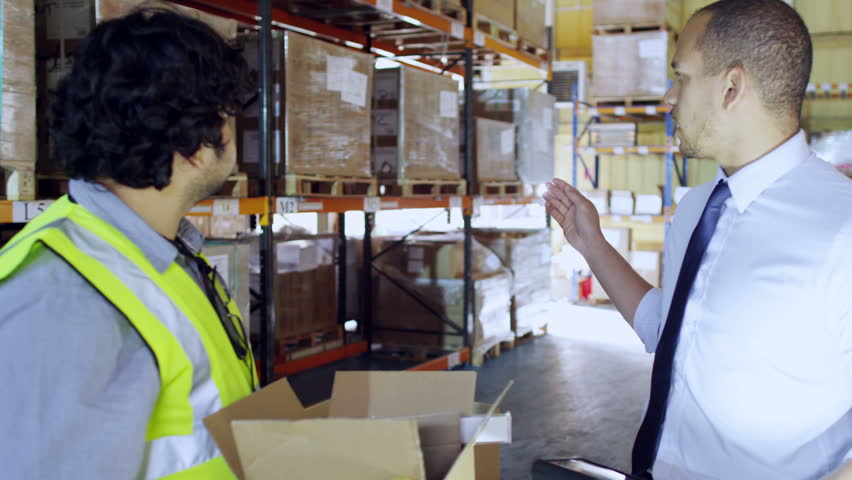 A young man is working in a warehouse, dressed in a shirt and tie and he is working on a digital tablet. He issues instructions to another male worker and another employee is working in the background