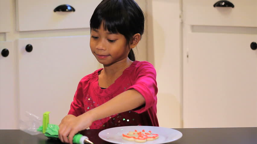 A cute little 6 year old Asian girl adds green icing to her yummy Christmas cookie.