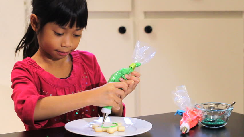 A cute little six year old Asian girl adds green icing to her yummy Christmas cookie.