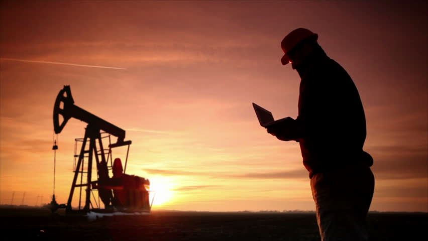 Oil Industry Pump jack with one oil worker who using a phone and typing on laptop.In a background are Silhouettes of one Pump jack at sunset light.
