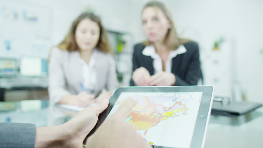 Two women and one man in business dress are having a meeting. The man's hands are all that we can see as he uses a digital tablet to flick through various graphs and statistics.