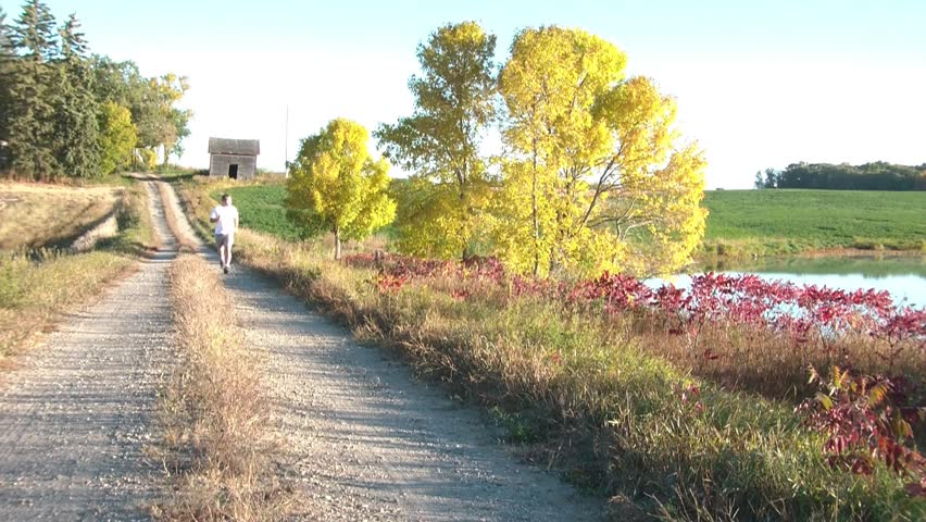 Guy jogs in autumn towards camera up a dirt road with scenic fall foliage, glassy lake, and a rustic rural structure in background.
