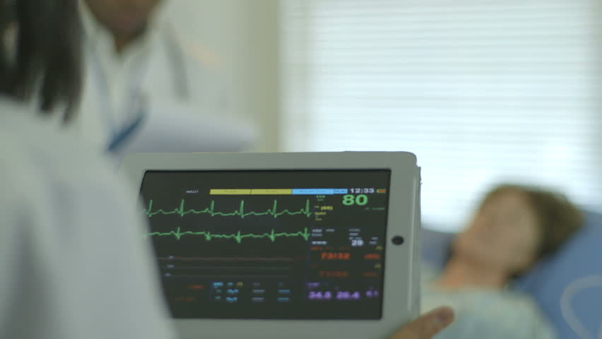 A doctor or nurse in a hospital setting looks at the vitals and other data being displayed on a portable patient monitor she is holding.
