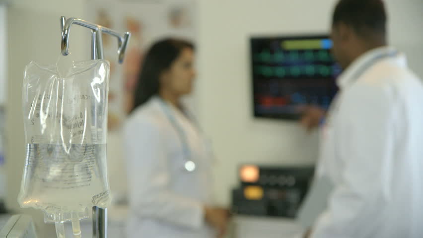 Focus moves from an IV bag solution in the foreground to two physicians consulting what they are studying on the screen of a patient monitoring device.