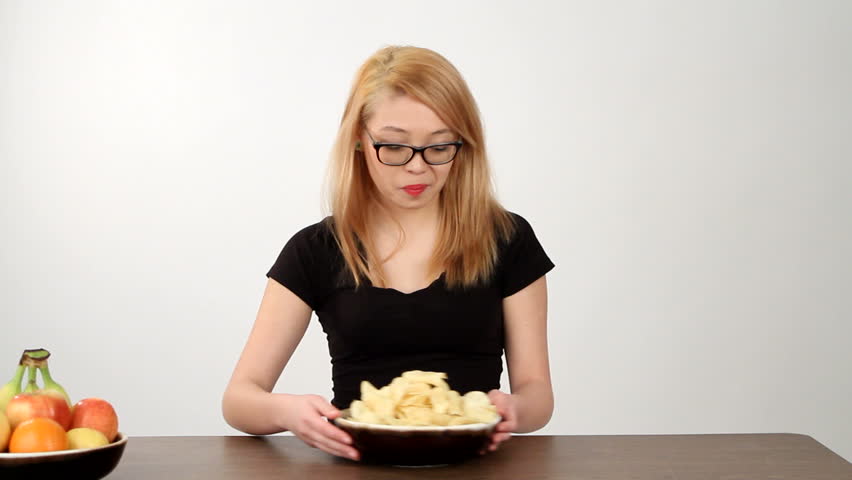 Teenager contemplates the choice of eating either some potato chips, junk food, or fruit, nutritious food, and chooses an junk food.
