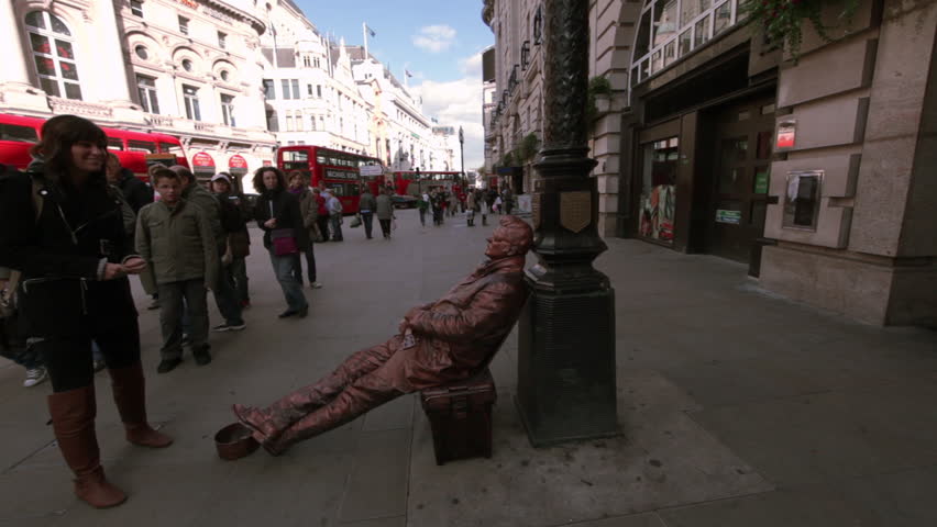 LONDON - OCTOBER 7, 2011: A lady putting money into the hat of a street performer sitting on a bench imitating a statue with red double decker buses at Piccadilly 