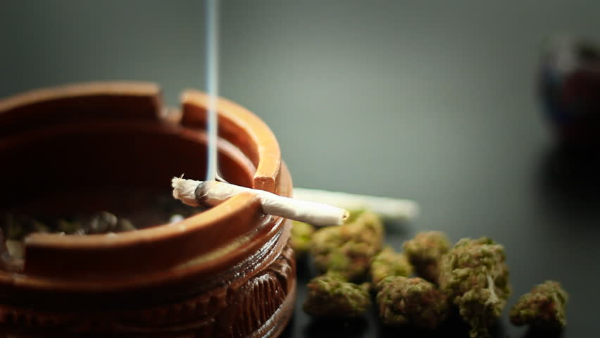 Marijuana Joint Burning 1. A marijuana filled joint burning on the edge of a wooden ashtray. More marijuana to the side.