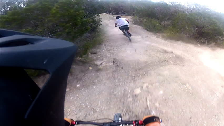 A mountain biker rides down a back country dirt path during the day while wearing a camera on his helmet. pov.