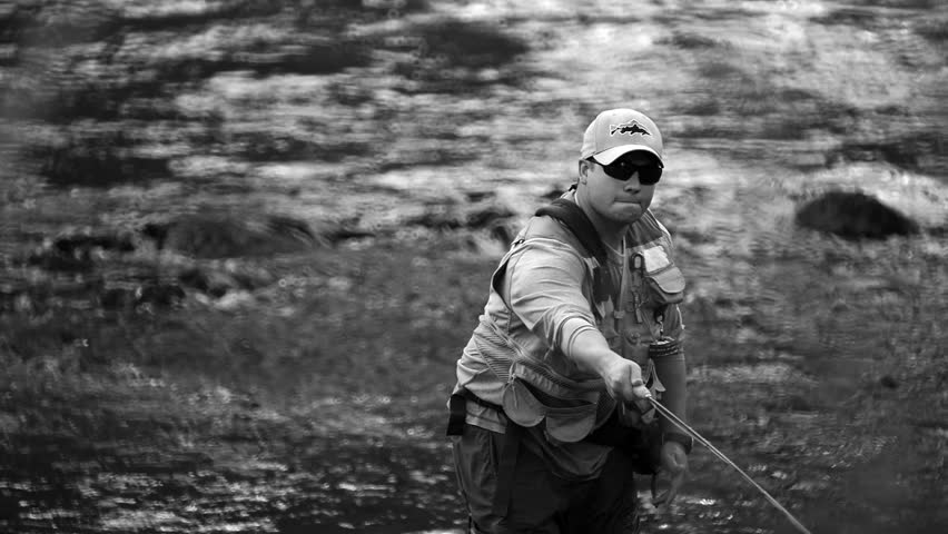 A man in waterproof overalls stands in a river and casts his lure with his rod while he spends his day fly fishing.