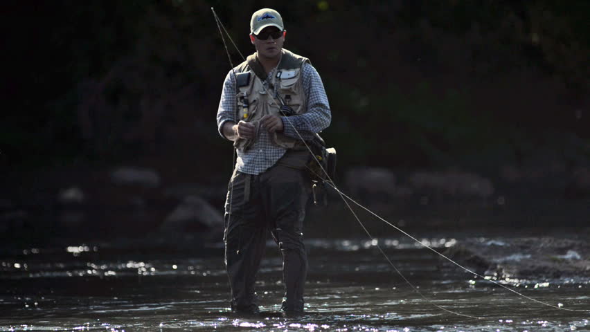 A man in waterproof overalls stands in a river and casts his lure with his rod while he spends his day fly fishing.
