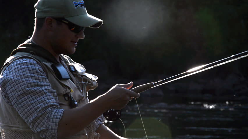A man in waterproof overalls stands in a river and casts his lure with his rod while he spends his day fly fishing.