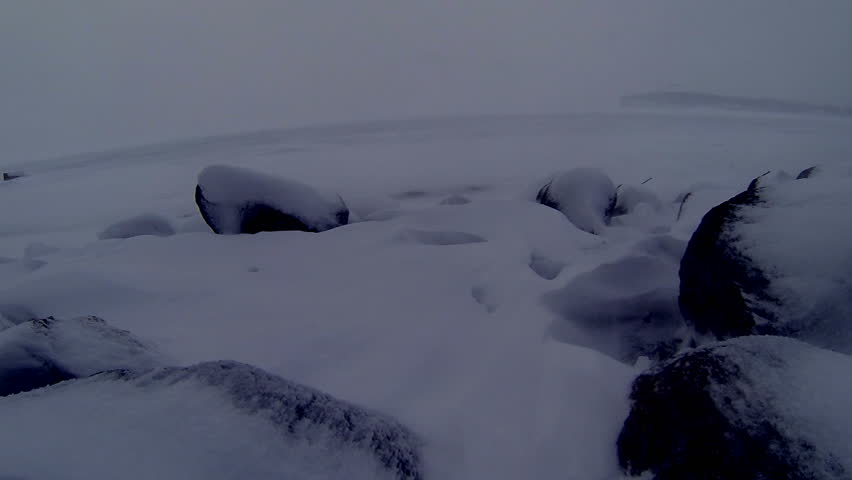 Snow blows towards the camera from across a wintry lake over rocks in the dim light of dusk. Slow motion video, shot at 60fps and conformed to 24fps (40% speed). Looping video.